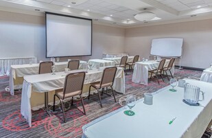Un salón de conferencias con varias mesas, sillas, una pantalla de proyector y una pizarra.