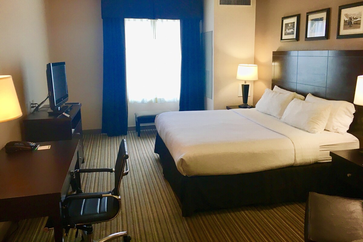 Quarto de hotel com cama, escrivaninha, cadeira, luminária e janela com cortinas.