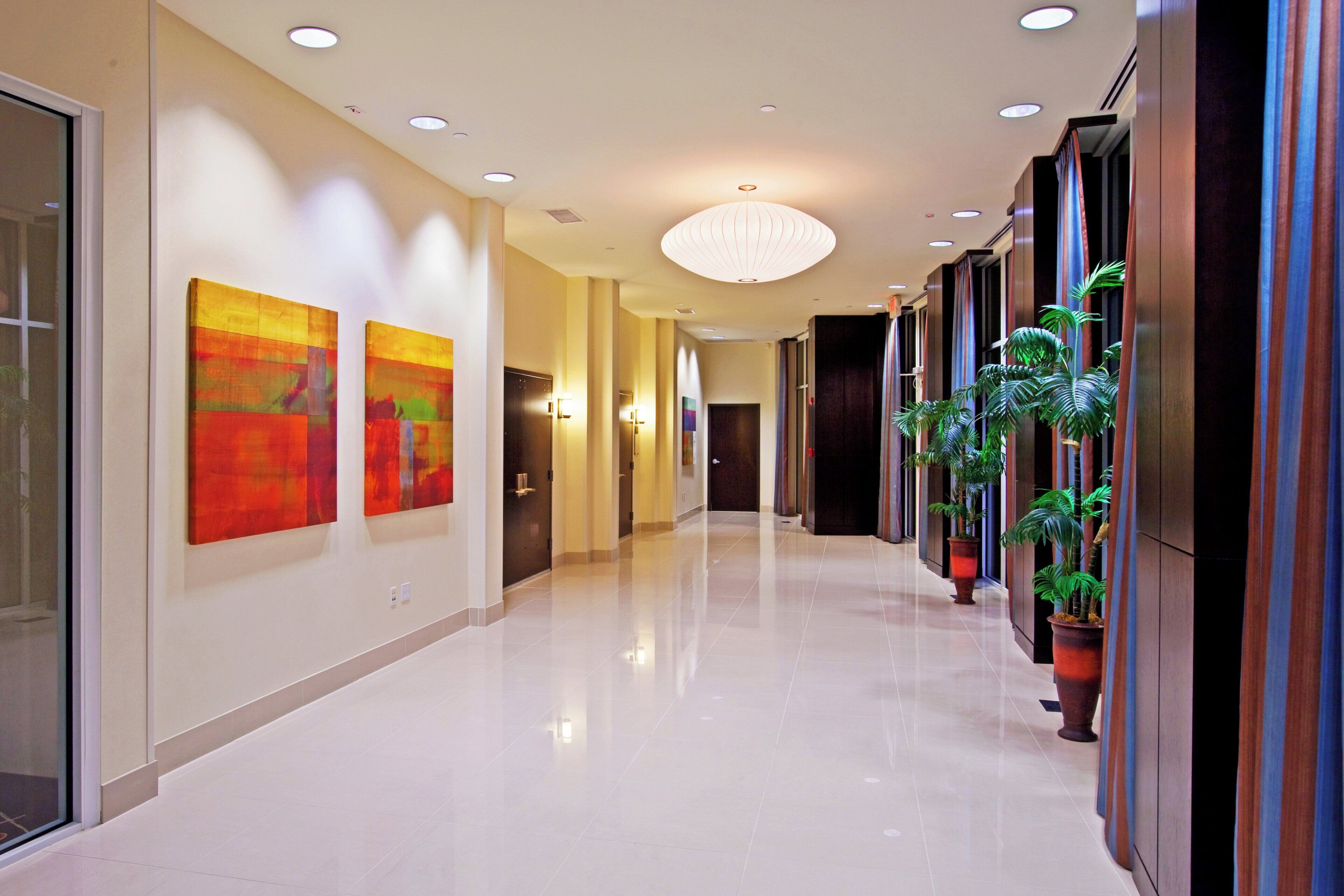 A hotel corridor with a polished floor, artwork on the walls, and potted plants along one side.