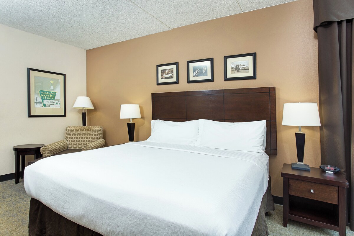 Quarto de hotel com uma cama grande, duas mesas de cabeceira com abajures, uma cadeira e quadros na parede.