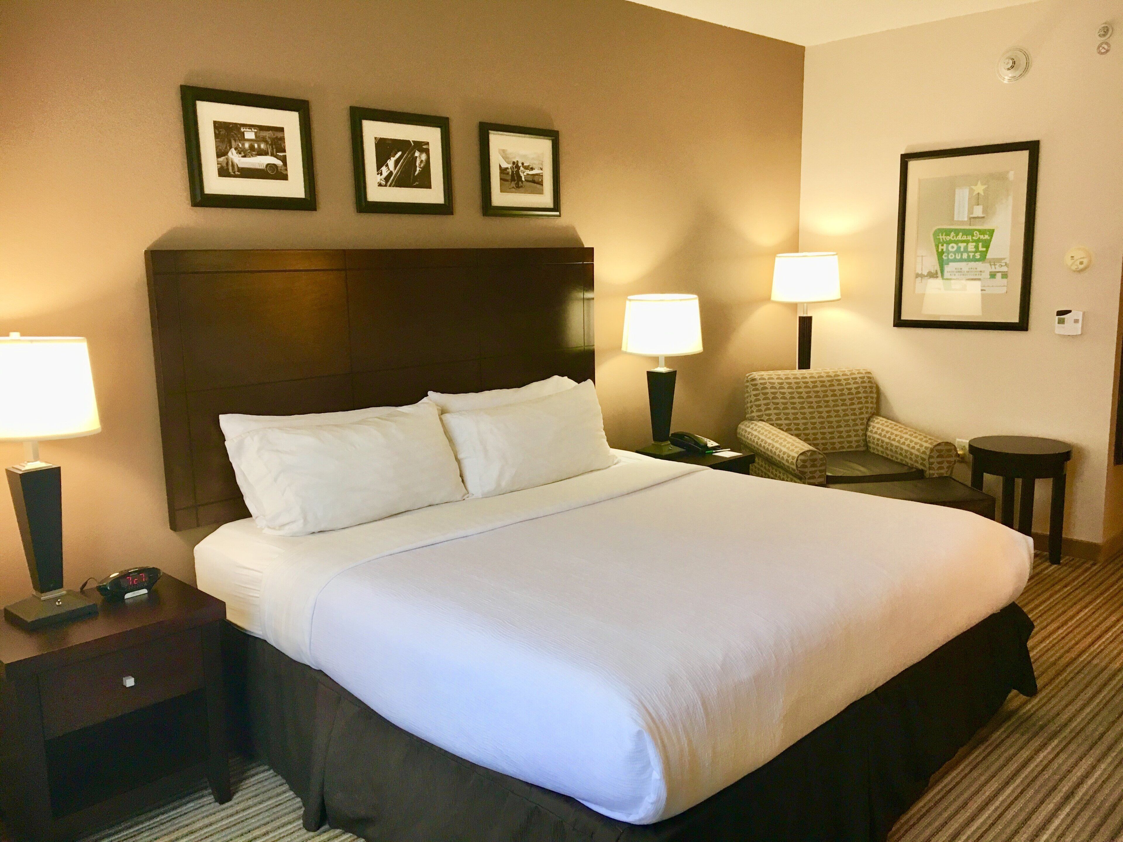 Una habitación de hotel con una cama grande, dos mesitas de noche con lámparas, una silla y cuadros en la pared.