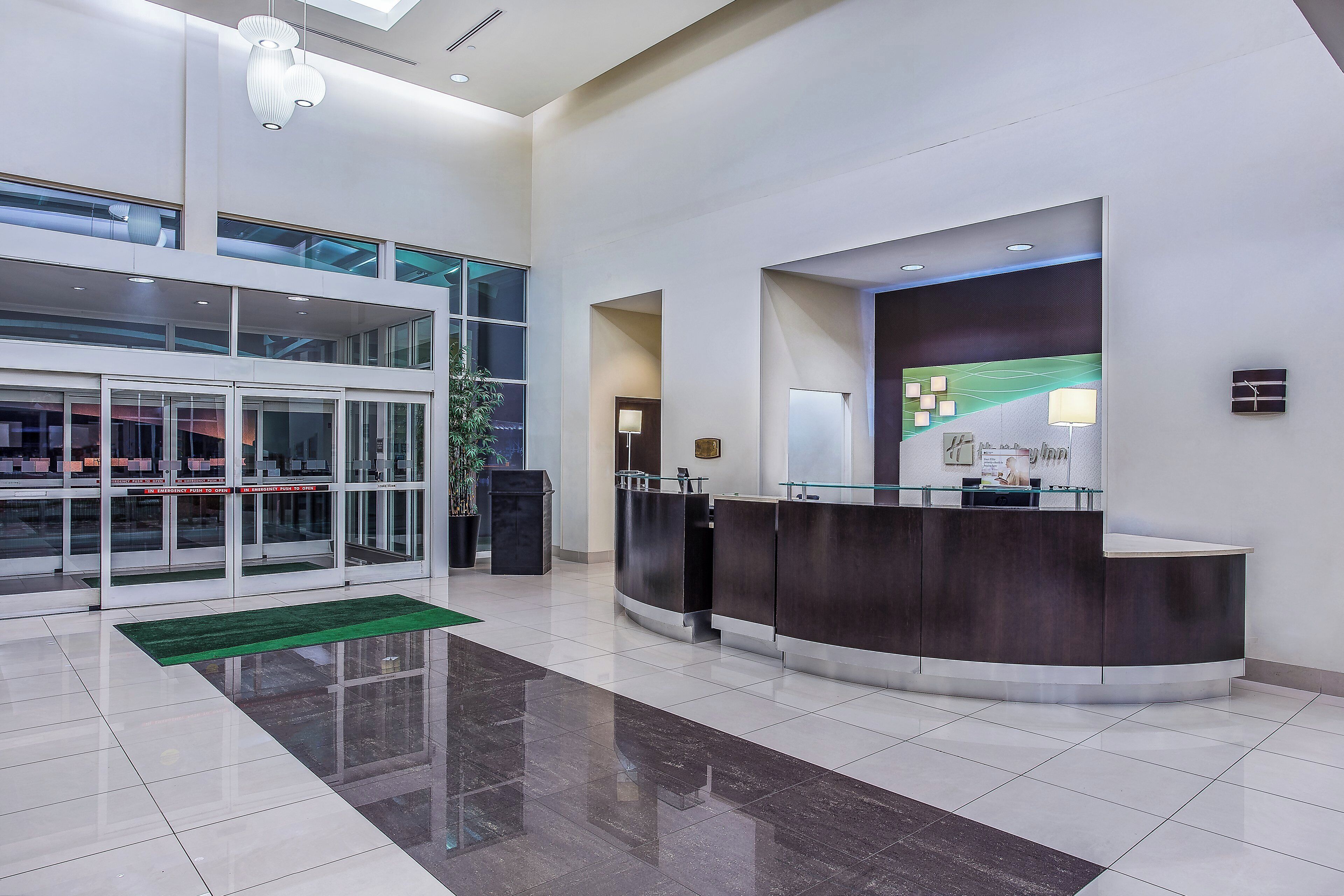 A modern hotel lobby with a reception desk, glass doors, and a green mat.