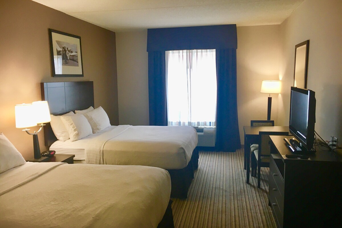 Quarto de hotel com duas camas, uma escrivaninha, televisão e janela com cortinas.