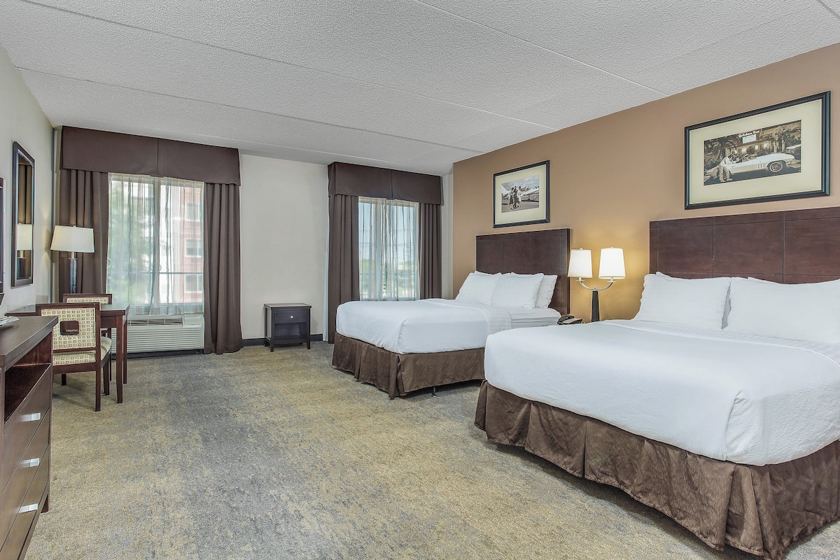 Quarto de hotel com duas camas, uma escrivaninha, uma cadeira, televisão e um quadro na parede.