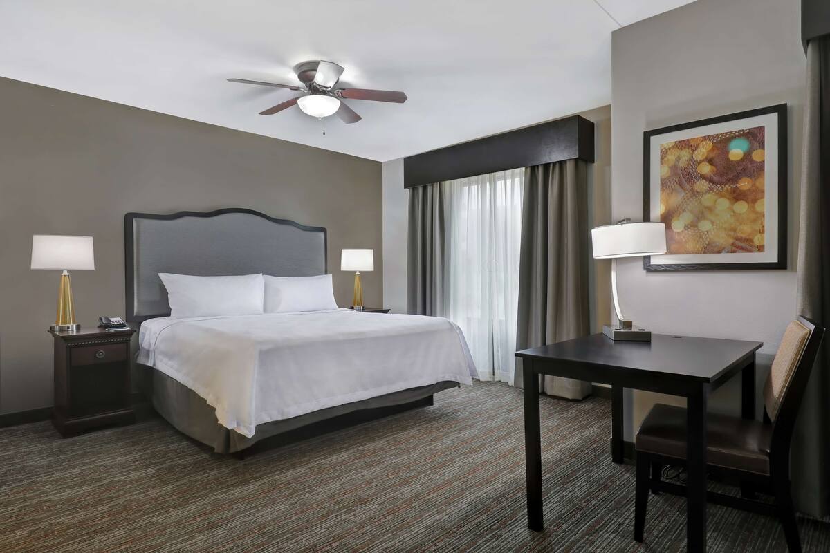Habitación de hotel con cama, mesitas de noche, escritorio, silla, ventilador de techo y un amplio ventanal con cortinas.