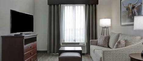 A hotel room with a flat-screen TV, a wooden dresser, a gray sofa, a small ottoman, a standing lamp, and a large window with curtains.