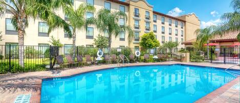 A swimming pool with a tiled deck, surrounded by palm trees and a hotel building with multiple windows and balconies.