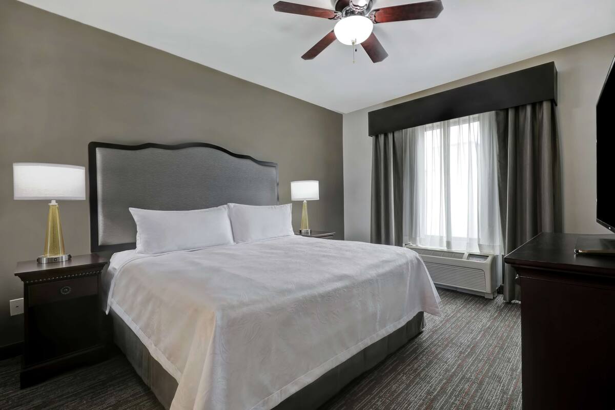 Una habitación de hotel con una cama grande, dos mesitas de noche con lámparas, un ventilador de techo, una ventana con cortinas y un televisor sobre un mueble.