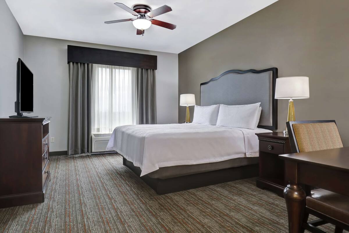 Habitación de hotel con cama, dos lámparas de noche, escritorio, silla, televisor, ventilador de techo y ventana con cortinas.
