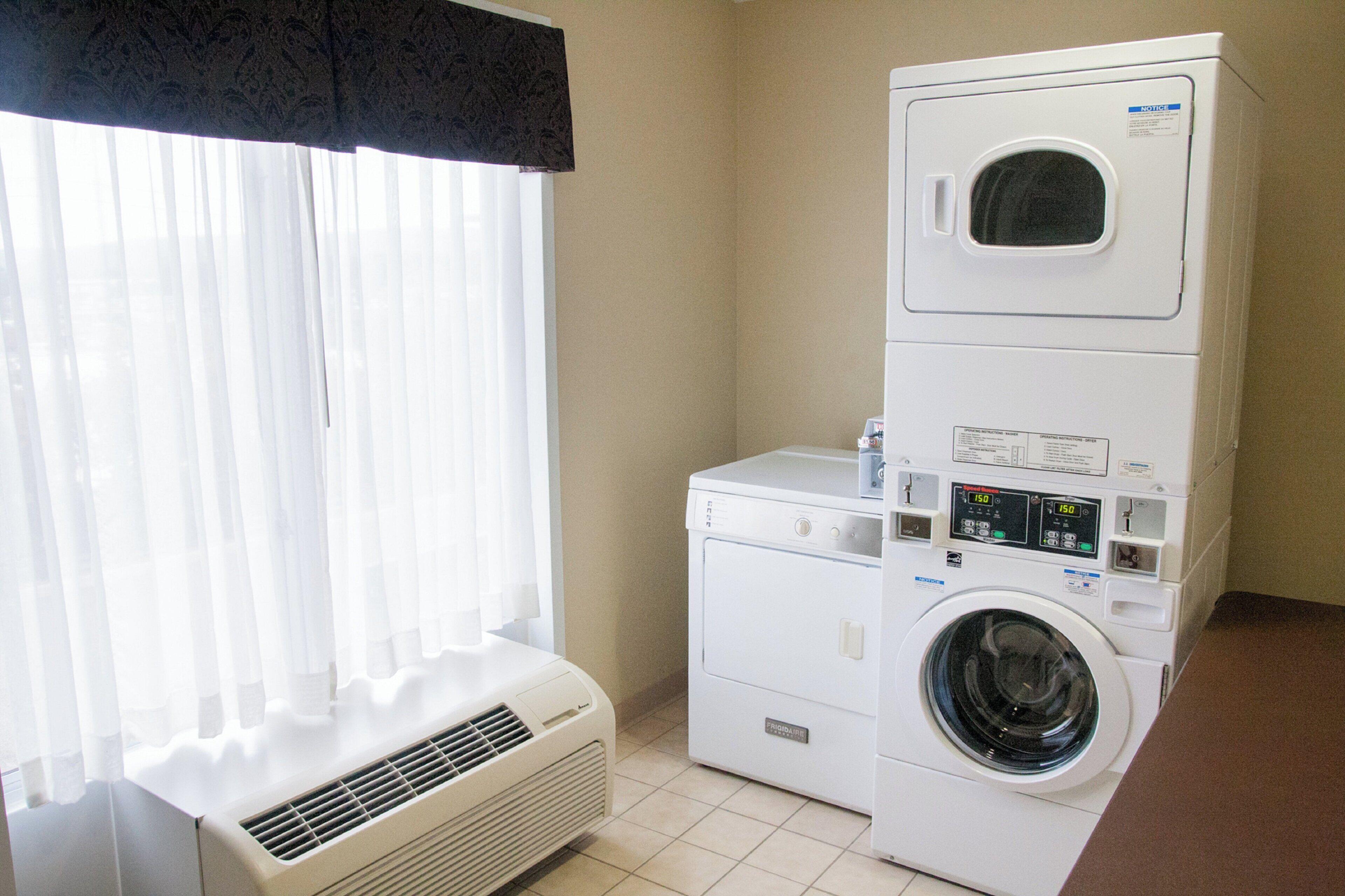A laundry area with stacked washers and dryers, a vent, and a window with sheer curtains.