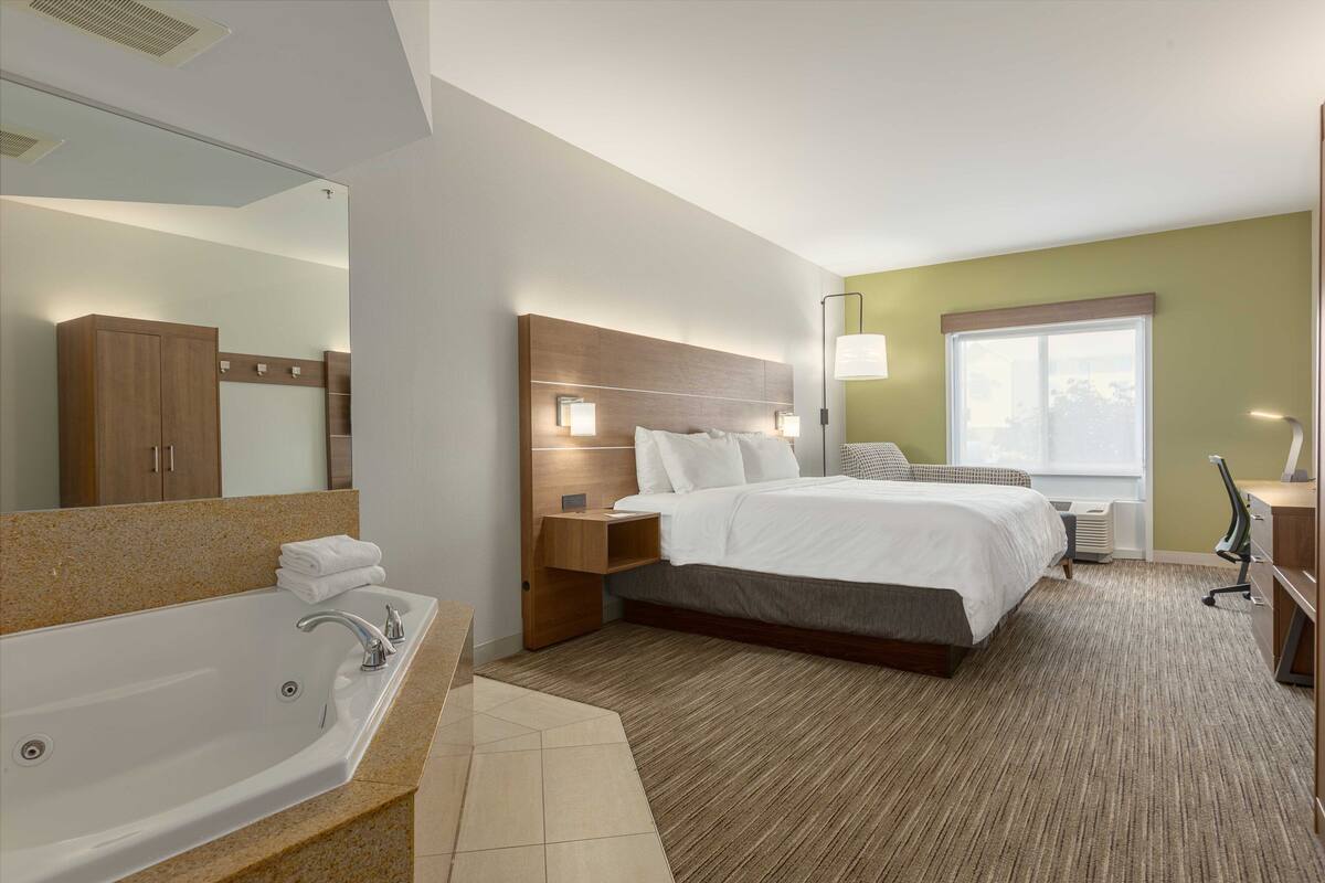 A modern hotel room with a large bed, a separate bathroom with a bathtub, a vanity with a mirror, and a large window providing natural light.
