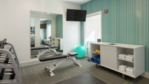 Un gimnasio con equipo de ejercicio, un espejo, un televisor y un estante con toallas.