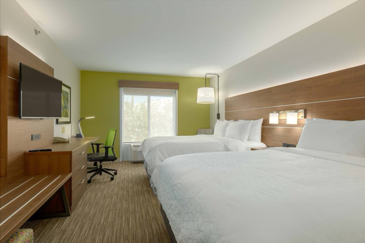 A hotel room with two beds, a desk with a chair, a television, and a window with a view of trees.