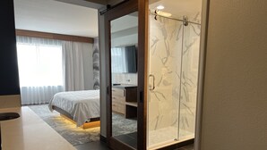 A modern hotel room with a bed, a desk, a shower with glass doors, and a view of the outdoors.
