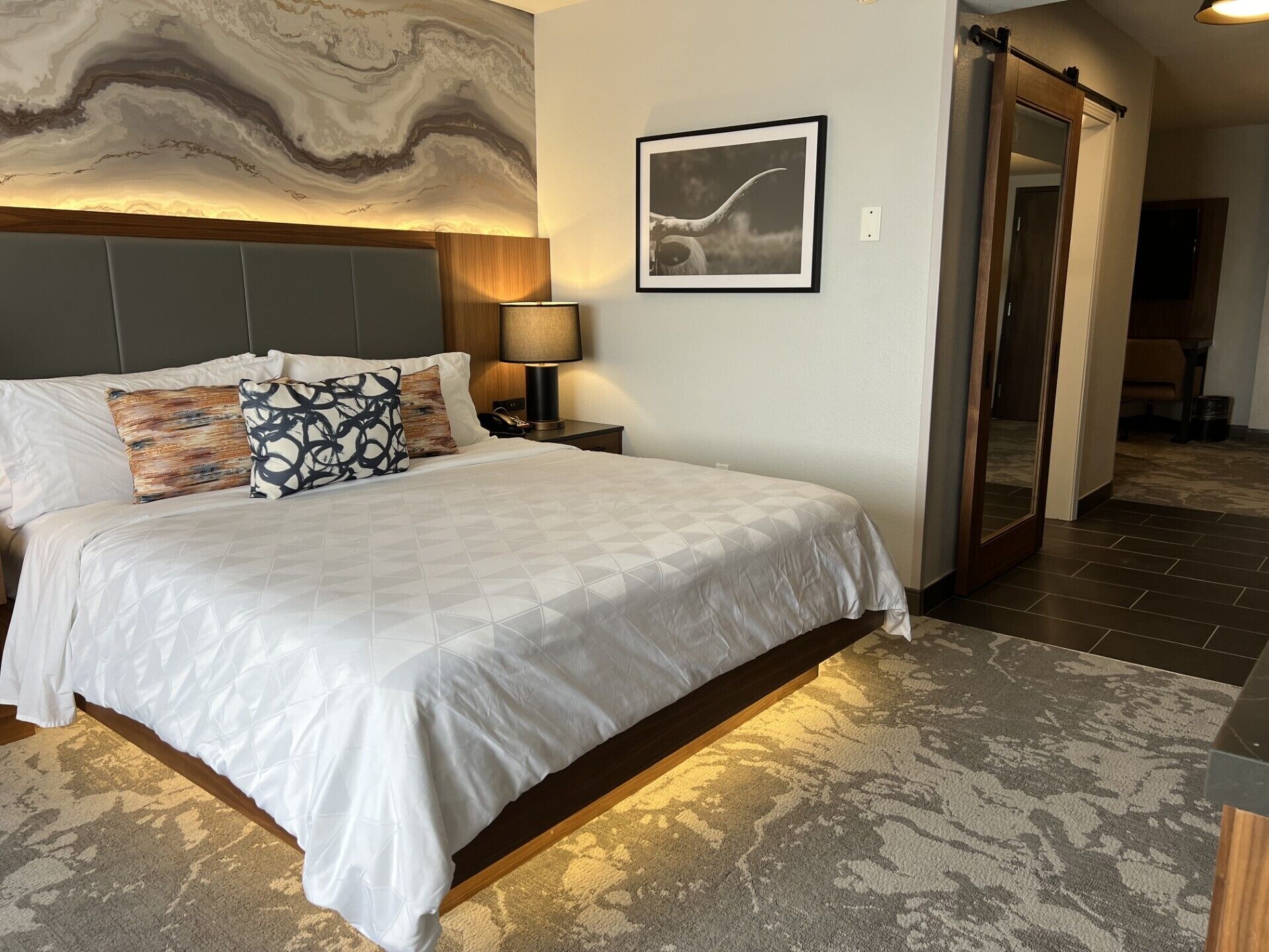 Une chambre d’hôtel avec un grand lit, une tête de lit, une table de chevet, une lampe, un tableau encadré et un miroir.