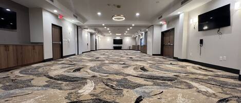A long, narrow hotel corridor with a patterned carpet, recessed lighting, and a flat-screen TV mounted on the wall.