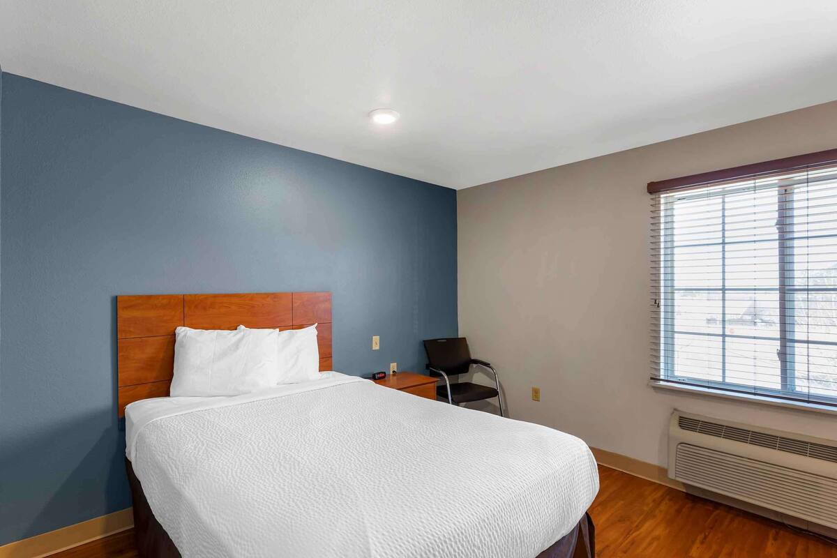 A hotel room with a bed, a chair, a nightstand, and a window with blinds.