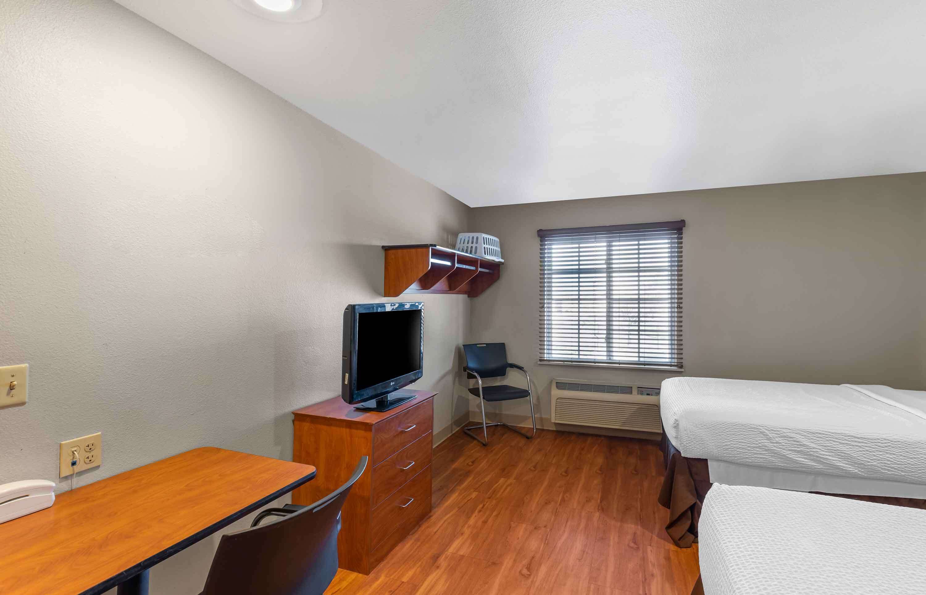 A small, well-lit room with a wooden desk, a chair, a television, a bed, and a window with blinds.