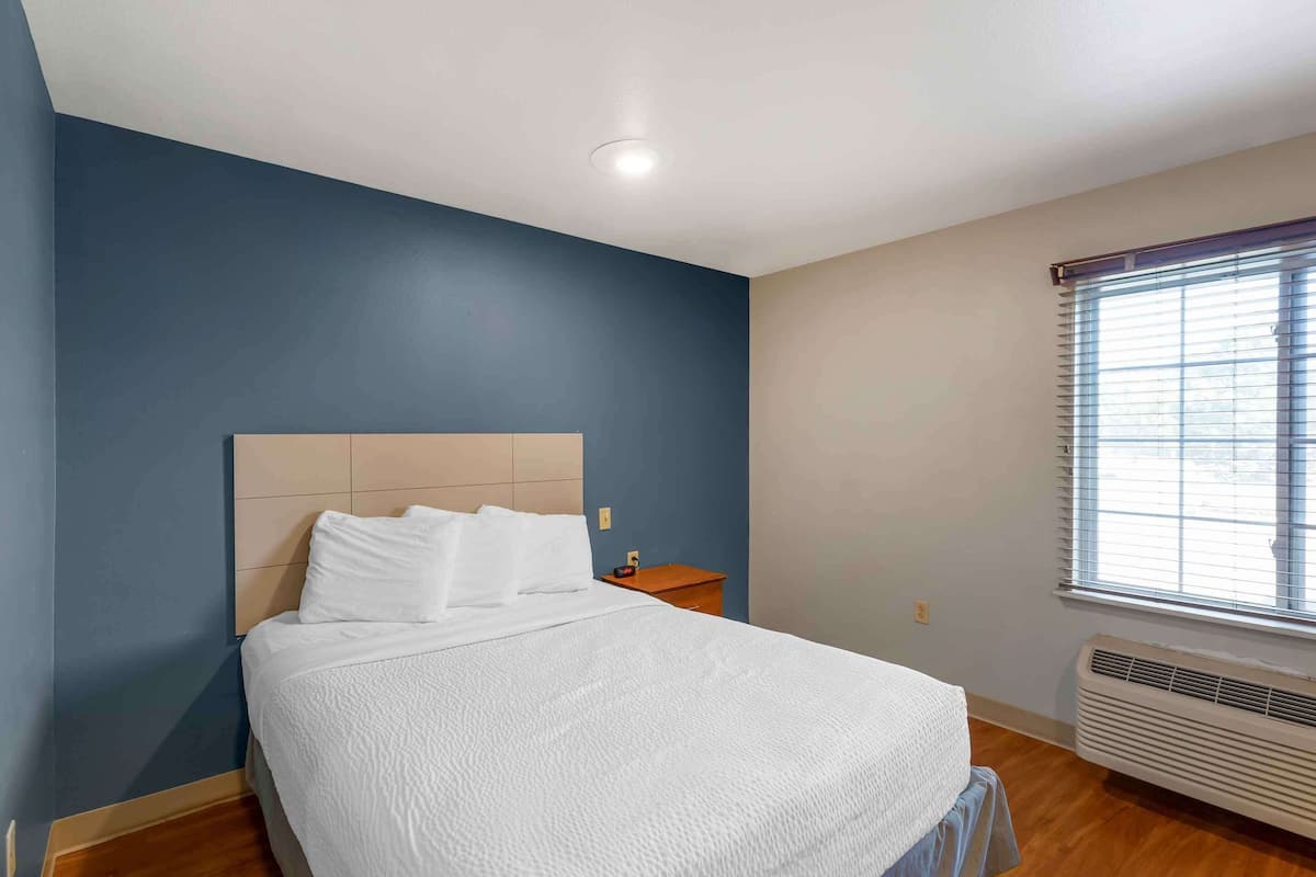 A neatly made bed in a room with a blue wall, a wooden floor, and a window with blinds.