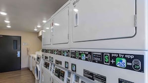 A row of white laundry machines with payment options via app, a door, and a light switch on the wall. - Extended Stay America Select Suites - Denver - Aurora (Aurora)