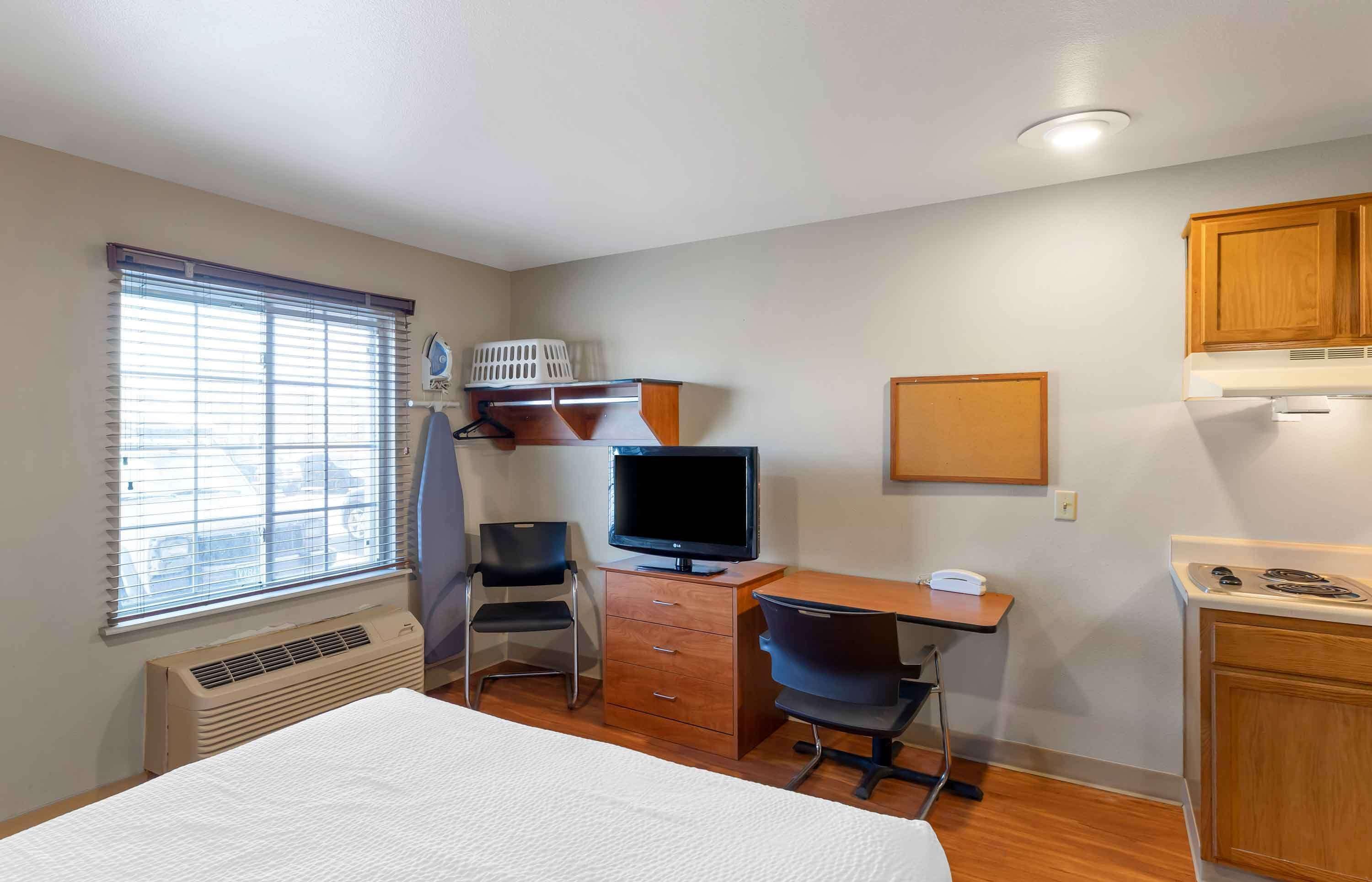 A hotel room with a bed, a desk with a computer, a chair, a television, a window with blinds, a heater, and wooden cabinets.