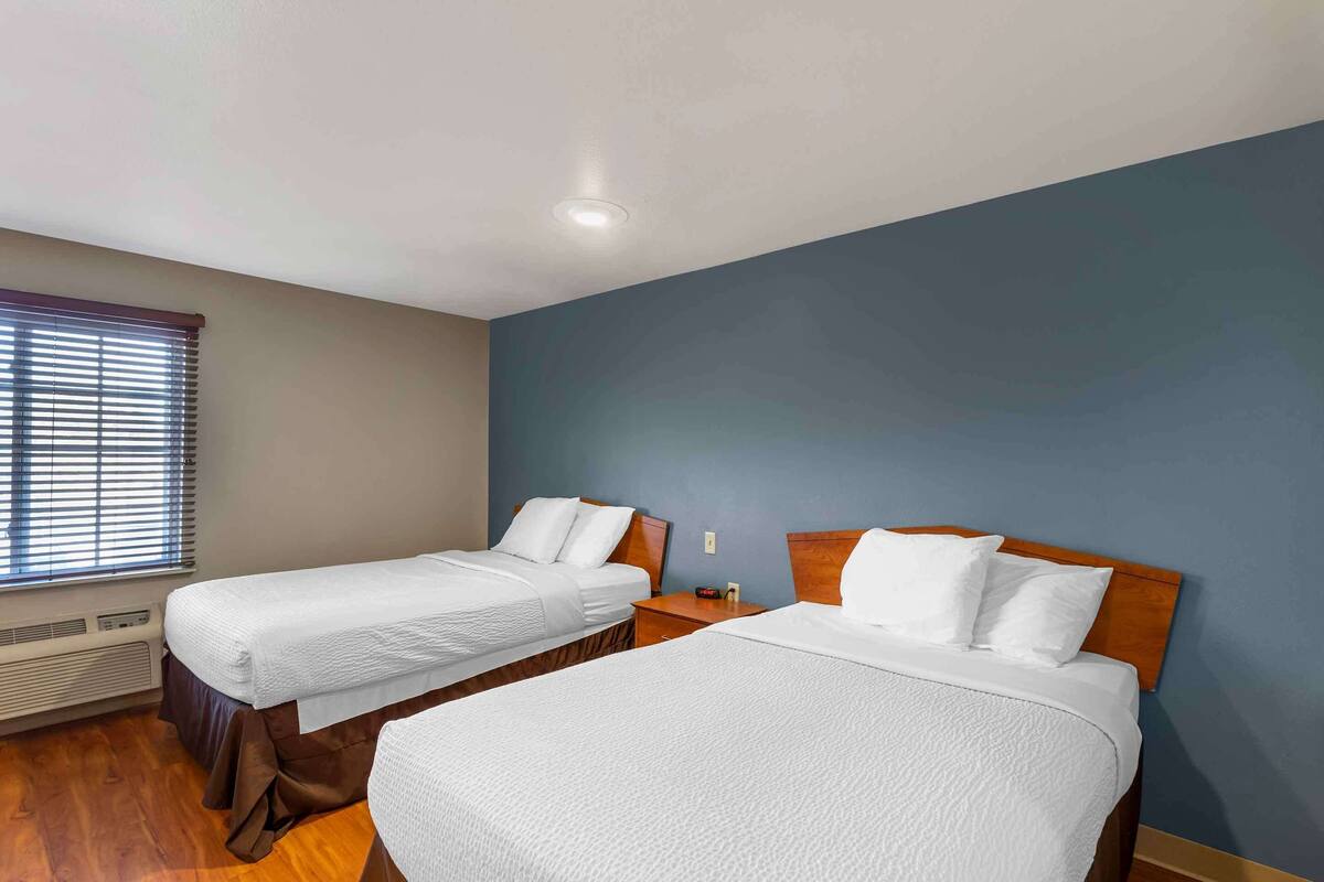 A hotel room with two single beds, a wooden headboard, a window with blinds, and a wall heater.