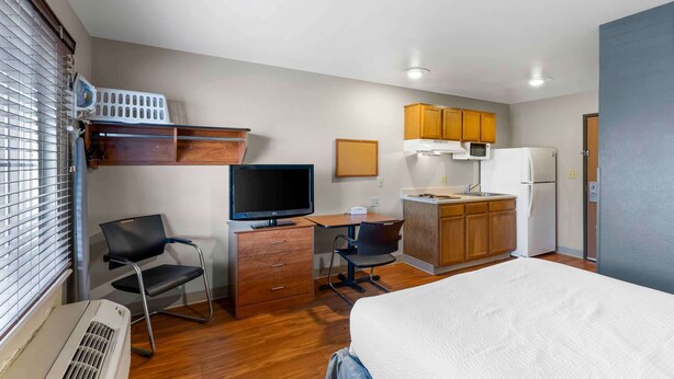 A compact hotel room with a bed, a desk with a computer, a TV, a kitchenette, and a window with blinds. - Extended Stay America Select Suites - Denver - Aurora (Aurora)