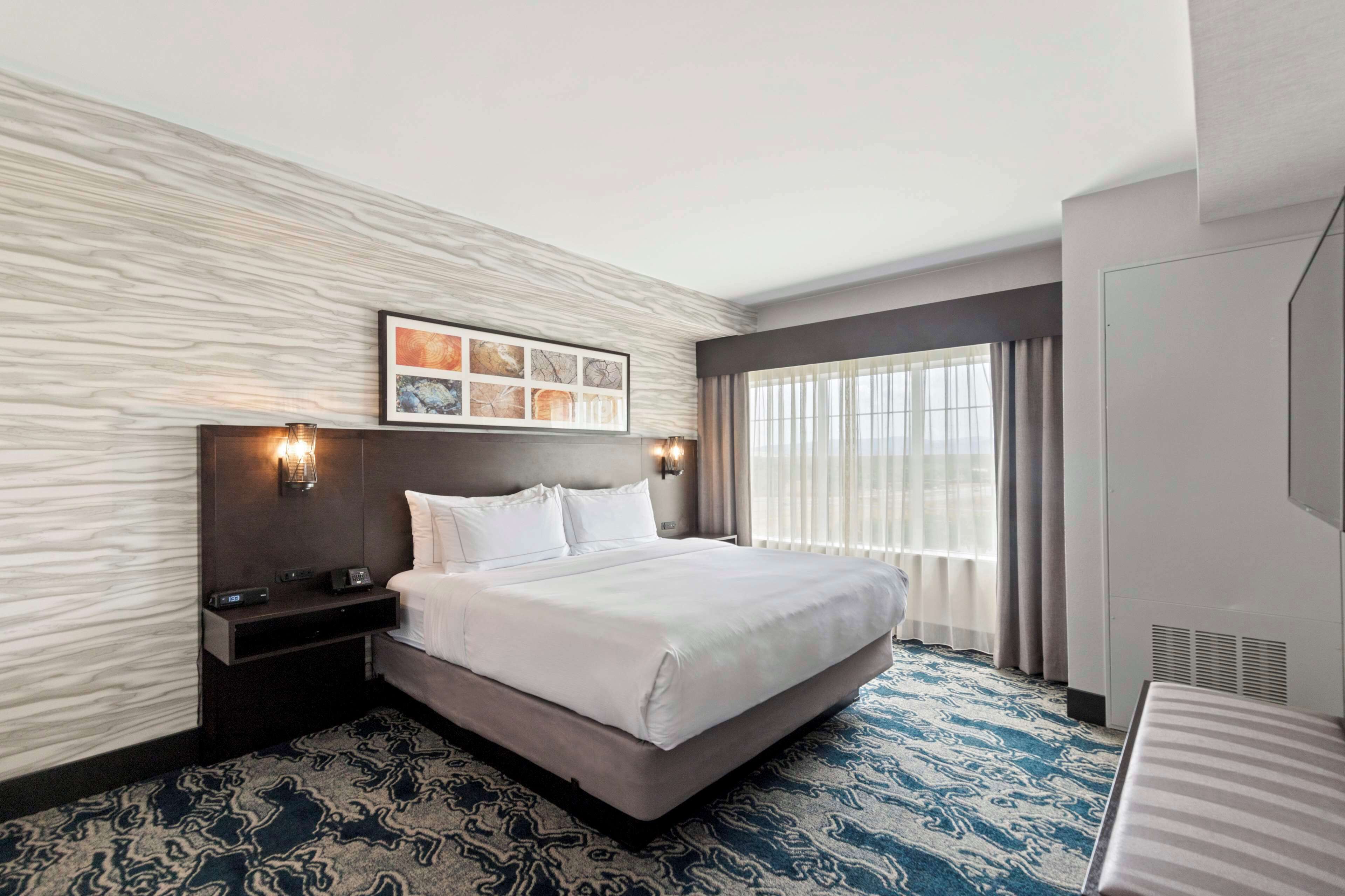 Ein Hotelzimmer mit einem großen Bett, einem Nachttisch, einem Wandbild und einem Fenster mit Vorhängen.