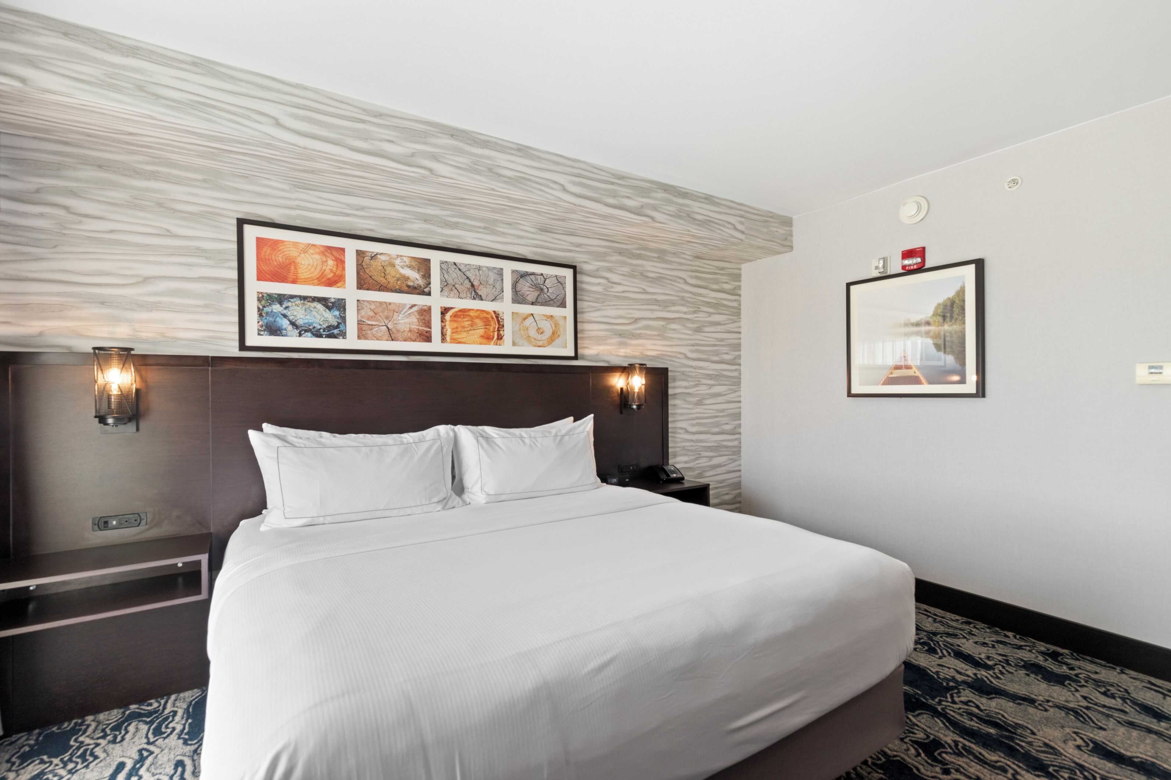Habitación de hotel con una cama grande, cabecera con obra de arte, mesita de noche y lámpara de pared.