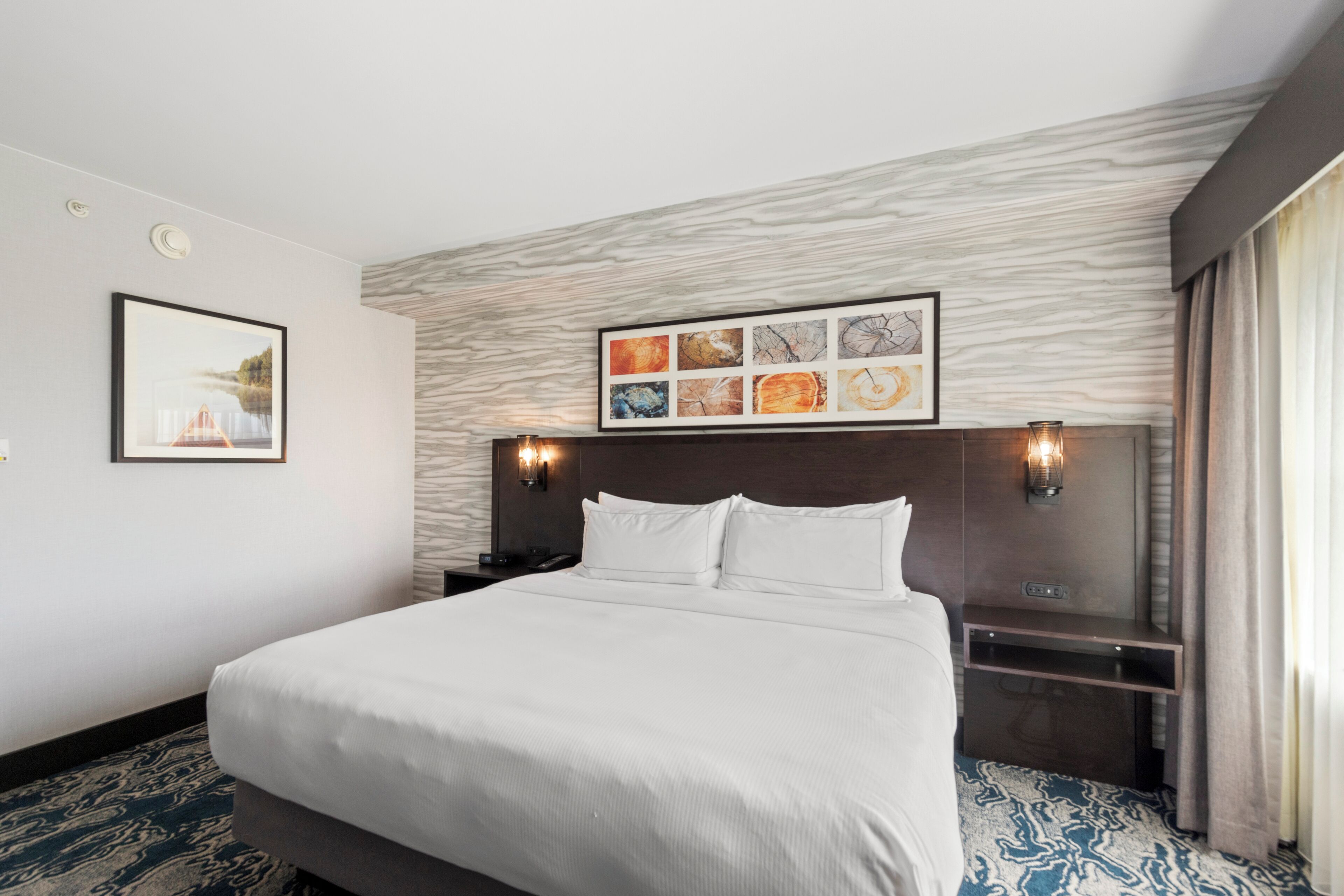 A hotel room with a large bed, a headboard with a light wood finish, a framed abstract art piece above the bed, and a nightstand with a lamp.