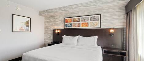Habitación de hotel con una cama grande, cabecera con acabado en madera clara, un cuadro abstracto enmarcado sobre la cama y una mesita de noche con una lámpara.