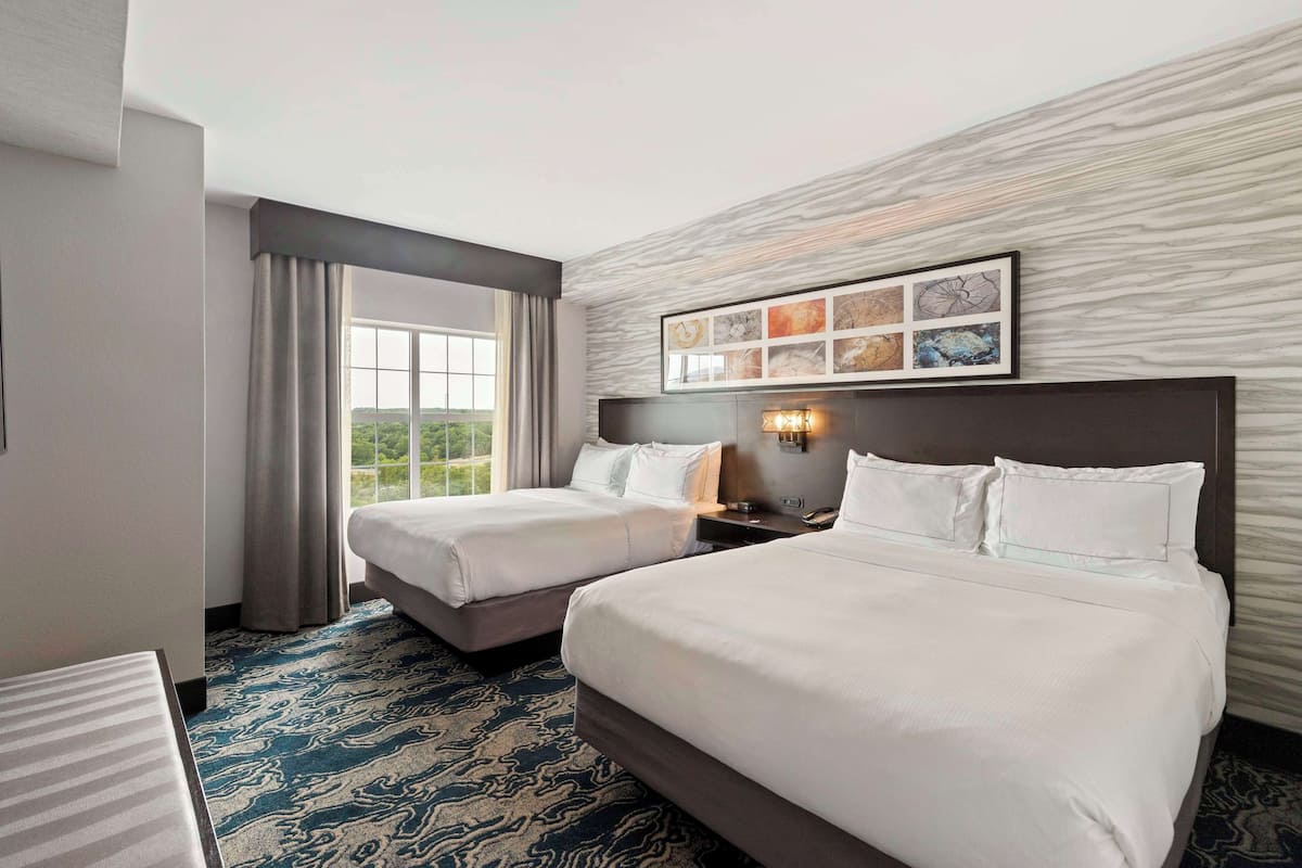 A hotel room with two beds, a large window with curtains, a marble-patterned wall, and a framed artwork above the beds.