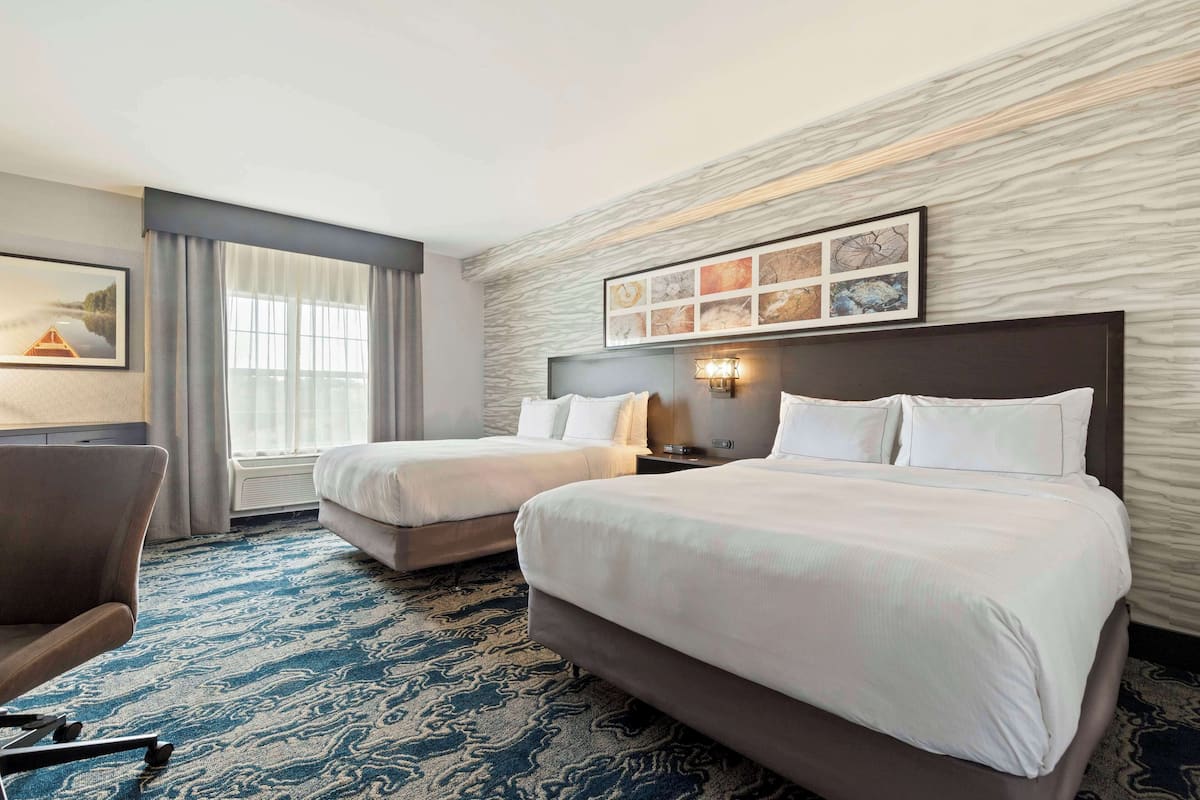 A hotel room with two beds, a desk, and a chair. There is a large window with curtains, a wall-mounted artwork, and a patterned carpet.