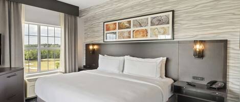 A hotel room with a large bed, a desk with a phone, and a window with a view of trees and a golf course.
