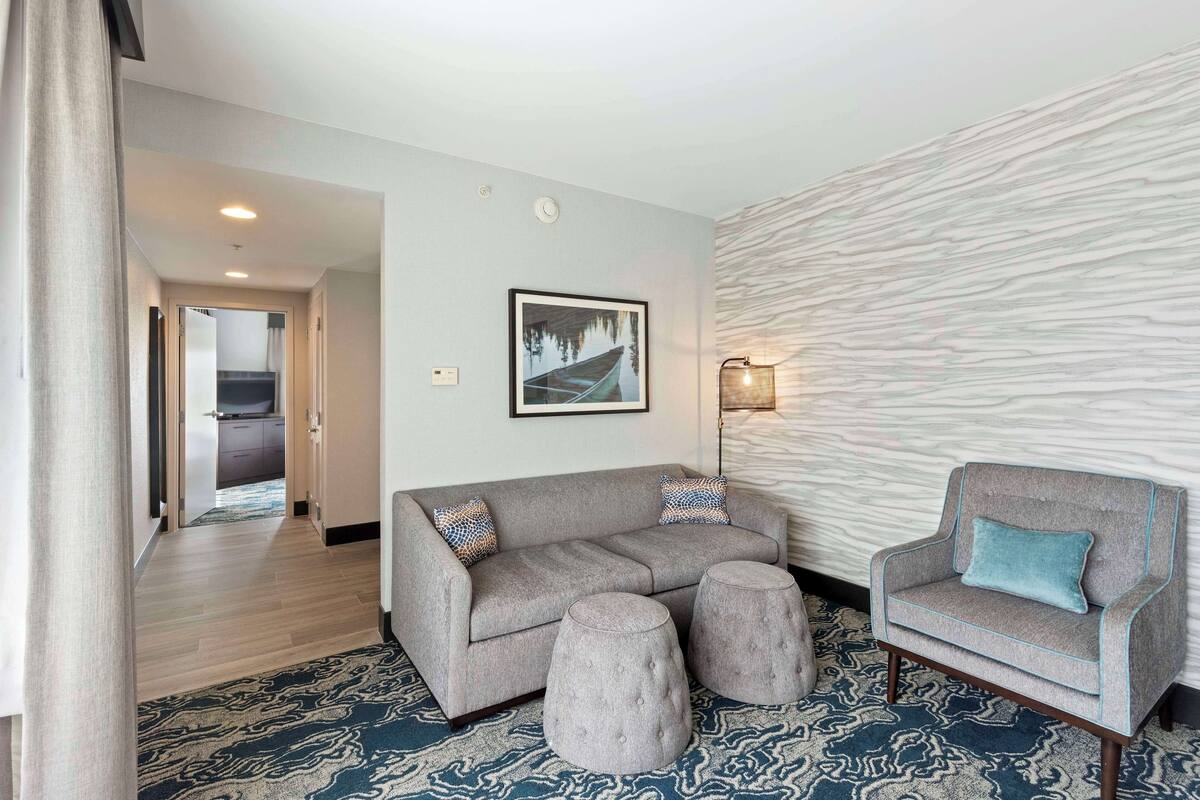 A modern living room with a grey sofa, ottomans, and a chair with a blue pillow. A framed picture of a lake and a boat is on the wall.