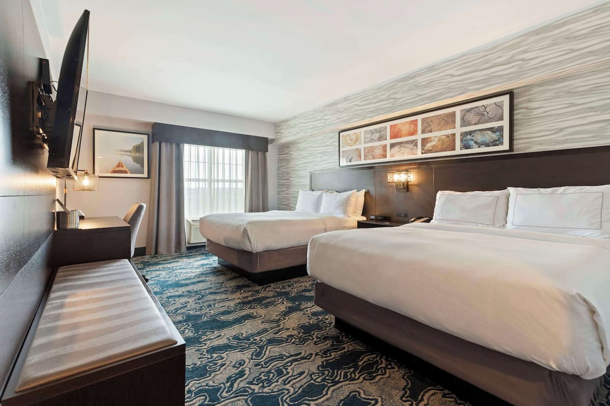 A hotel room with two beds, a desk, a chair, a TV, and artwork on the wall.