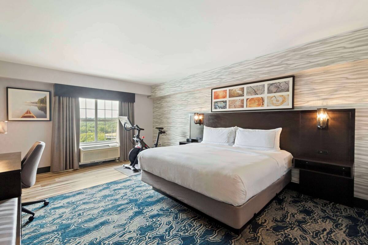 A hotel room with a large bed, a desk, a chair, a bike, and a view of the outdoors.