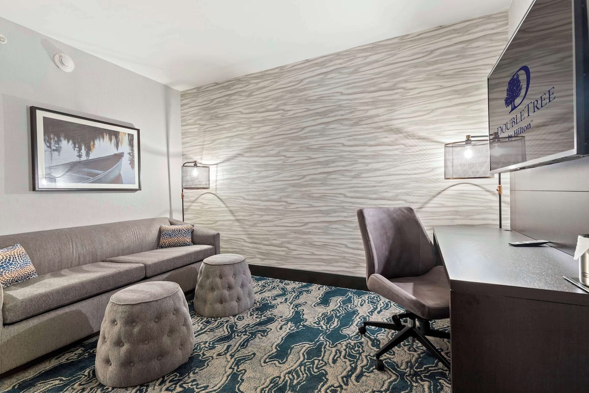 A modern hotel room with a grey sofa, two ottomans, a desk, and a wall-mounted lamp.