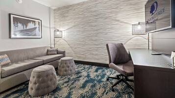 A modern hotel room with a grey sofa, two ottomans, a desk, and a wall-mounted lamp.