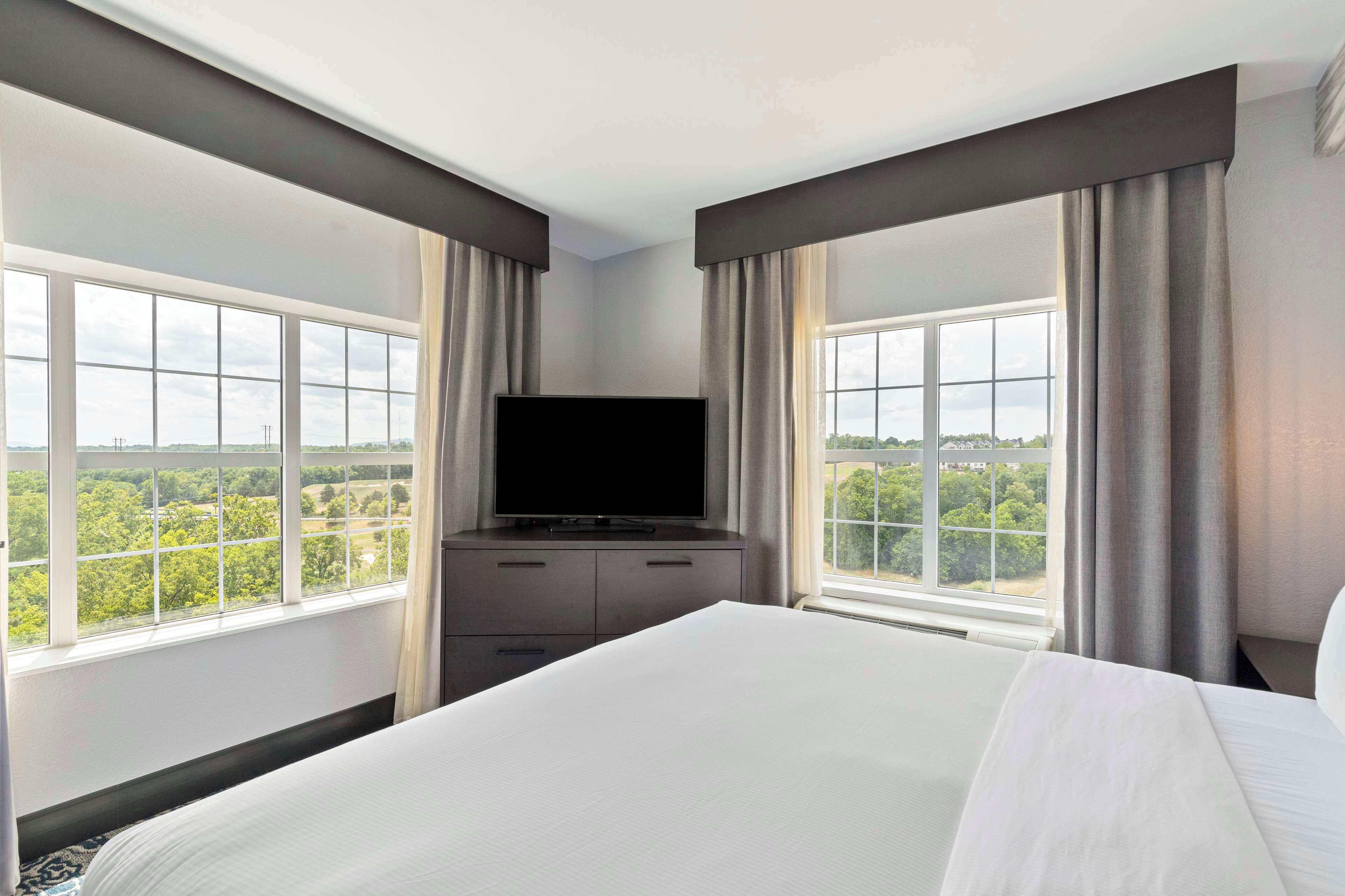 A hotel room with a large bed, a flat-screen TV, a dresser, and a view of a green landscape through large windows.
