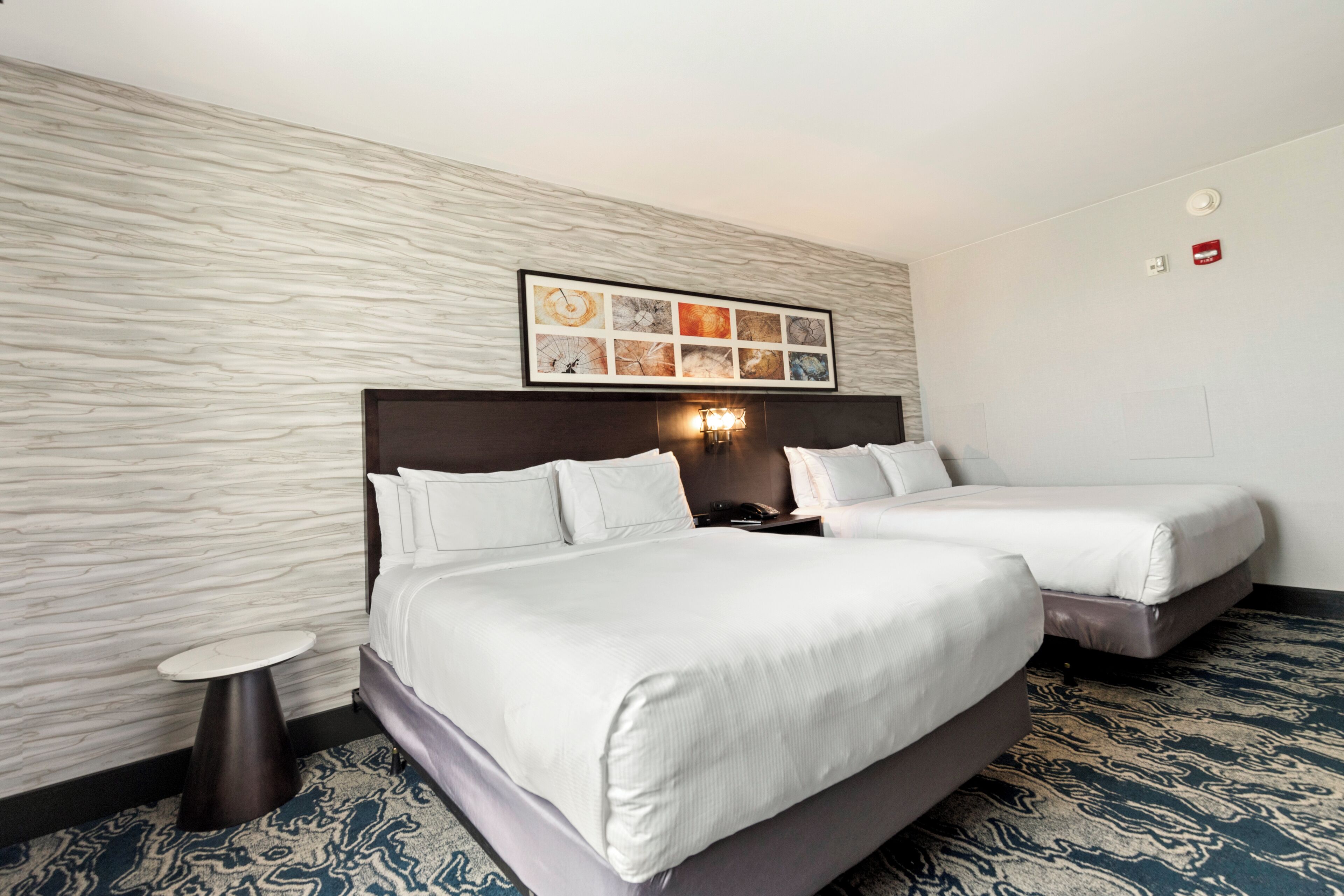 A hotel room with two beds, a small round table, a wall-mounted artwork, and a patterned carpet.