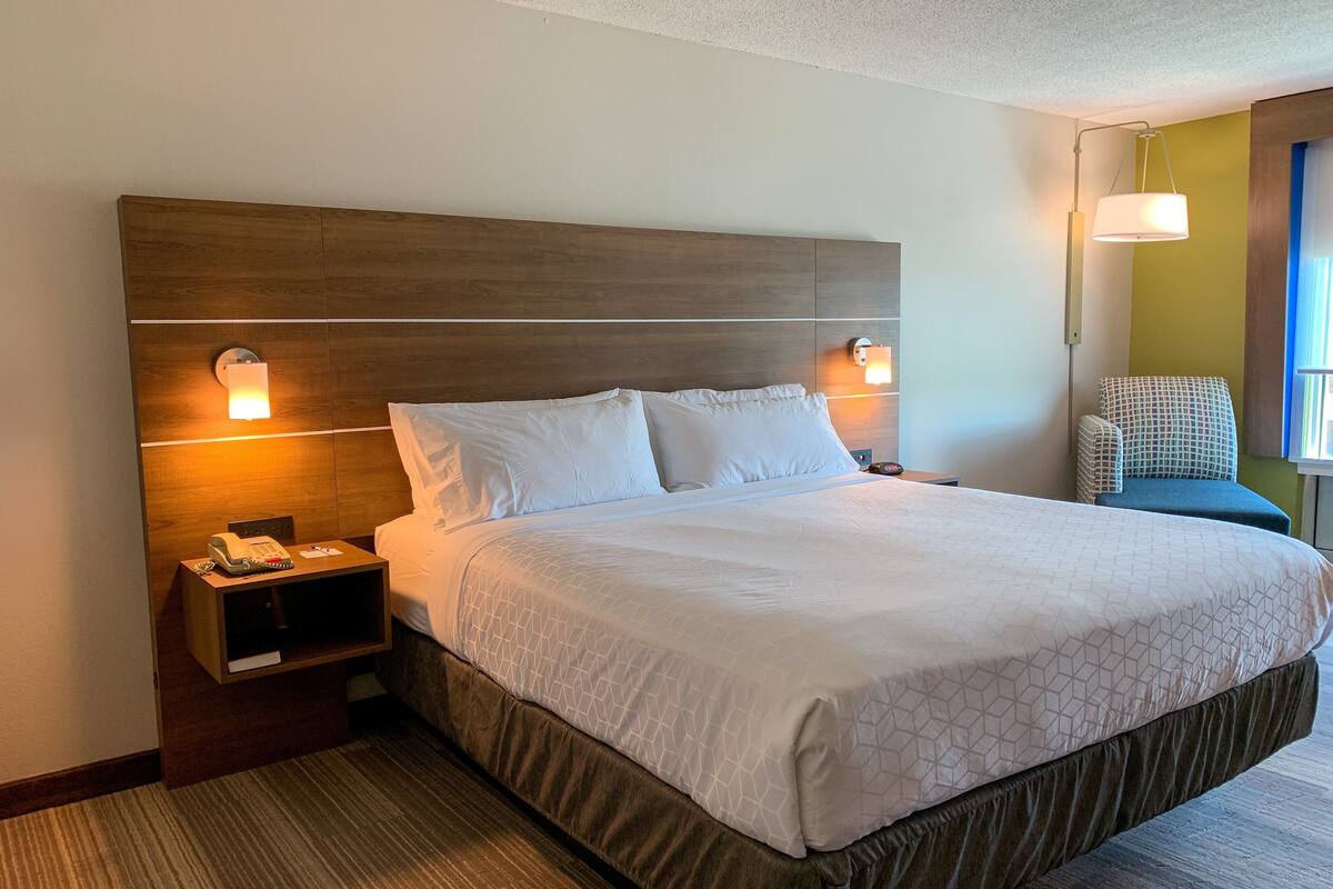 A hotel room with a large bed, two bedside lamps, a phone on the nightstand, and a chair with a patterned cushion.