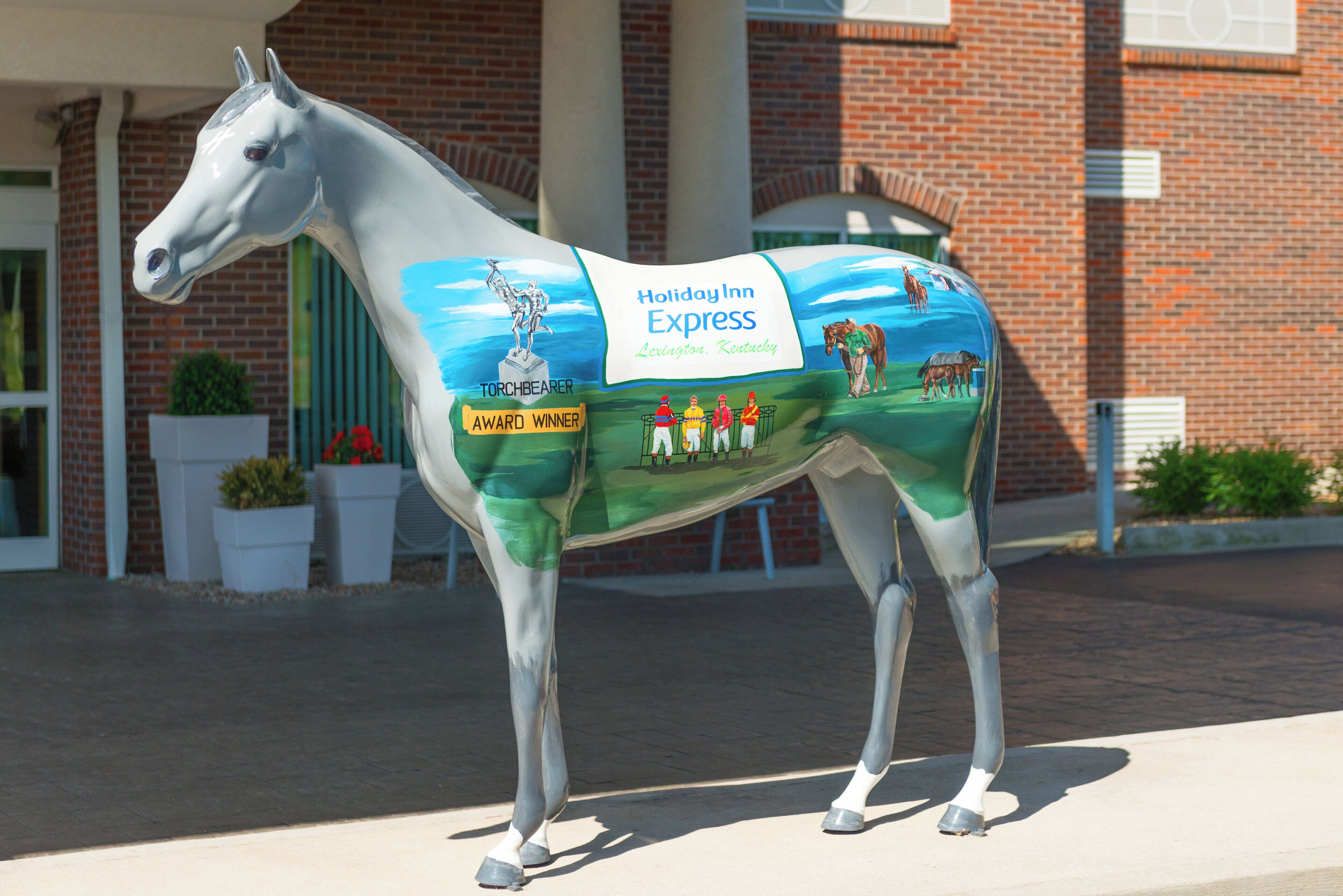 Une statue de cheval sur laquelle est peinte une publicité pour l’hôtel Holiday Inn Express de Lexington, dans le Kentucky. On y voit un lauréat du prix « Torchbearer Award » ainsi qu’un groupe de jockeys.