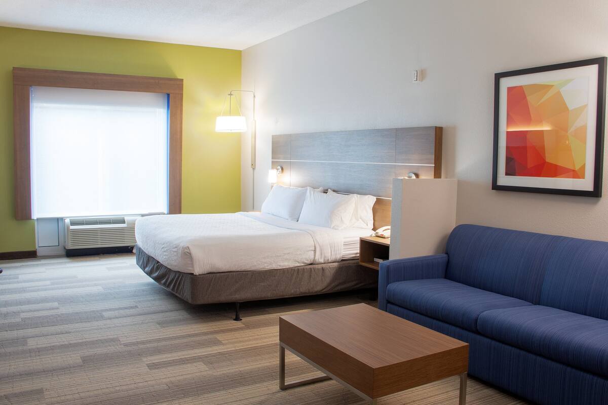 A hotel room with a bed, a blue sofa, a wooden coffee table, and a framed abstract painting on the wall.