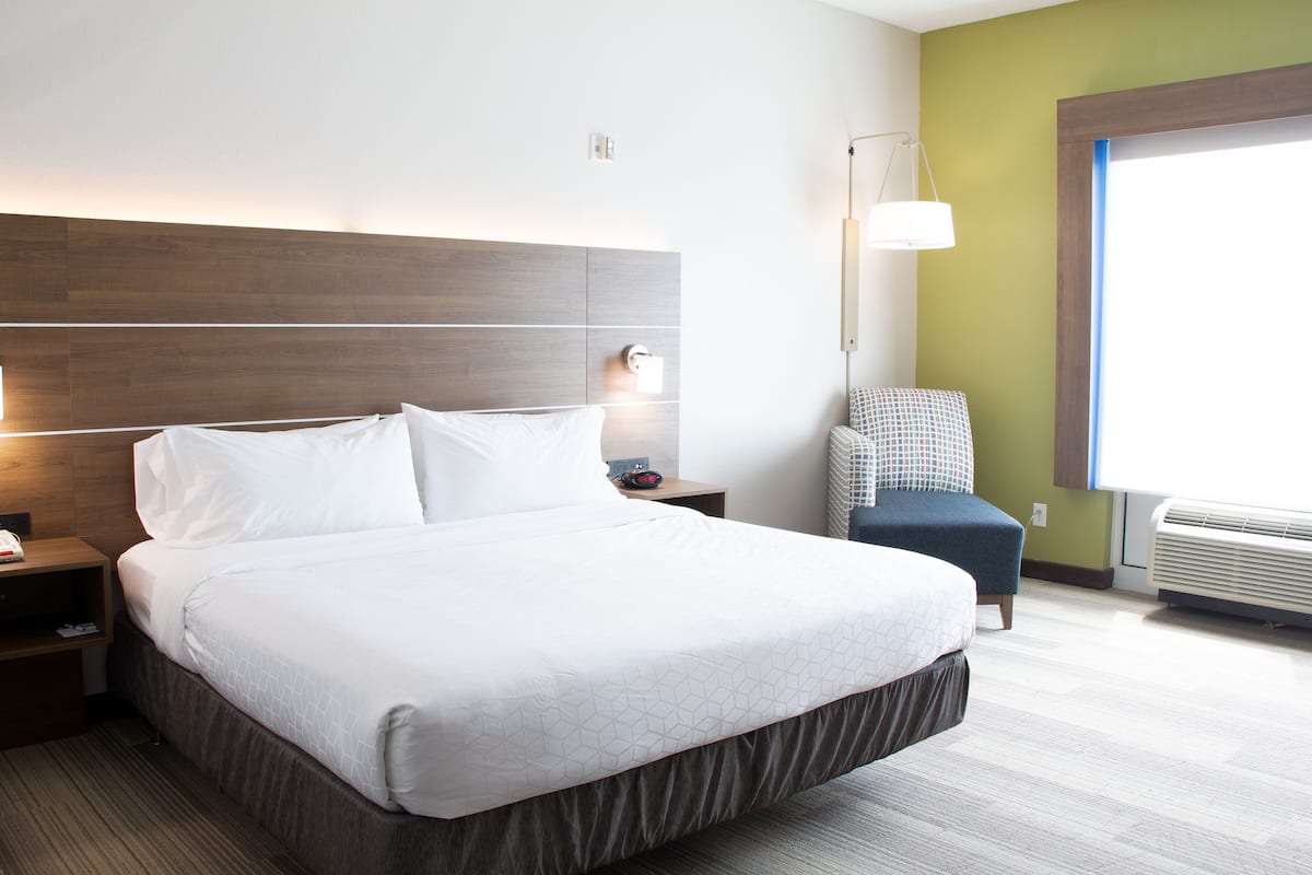 A hotel room with a large bed, a nightstand, a chair, a lamp, and a window with blinds.