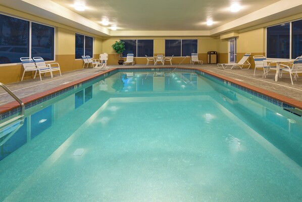 Una piscina interior rectangular con agua cristalina, rodeada de sillas reclinables blancas y una planta en maceta.
