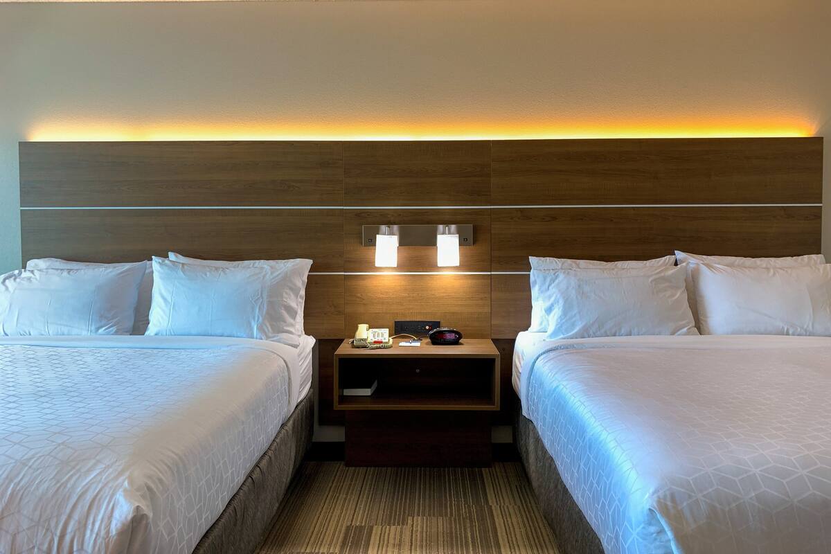 A hotel room with two beds, a nightstand with a phone and alarm clock, and a wall-mounted light fixture.