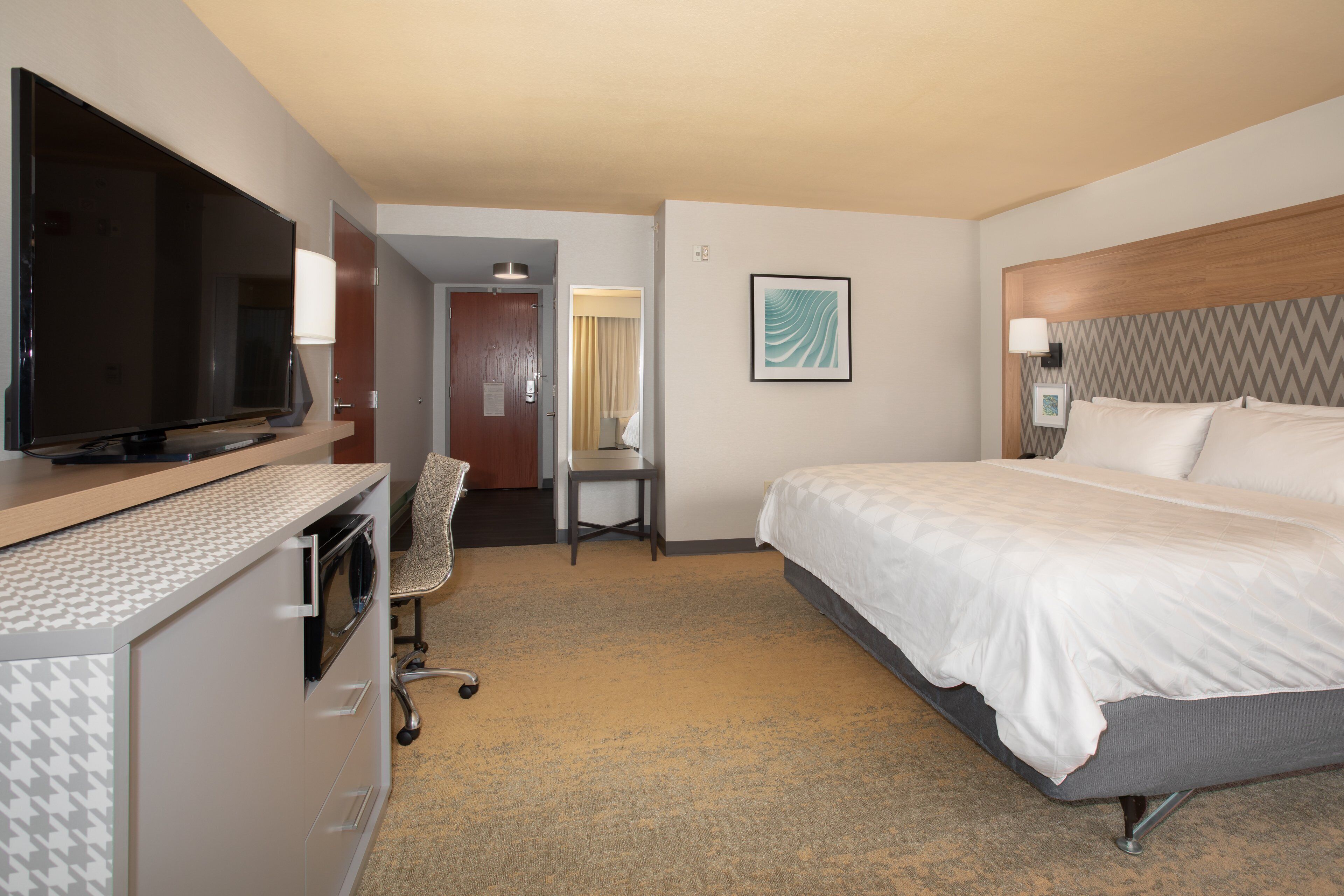 A hotel room with a bed, a desk with a chair, a television, and a framed artwork on the wall.
