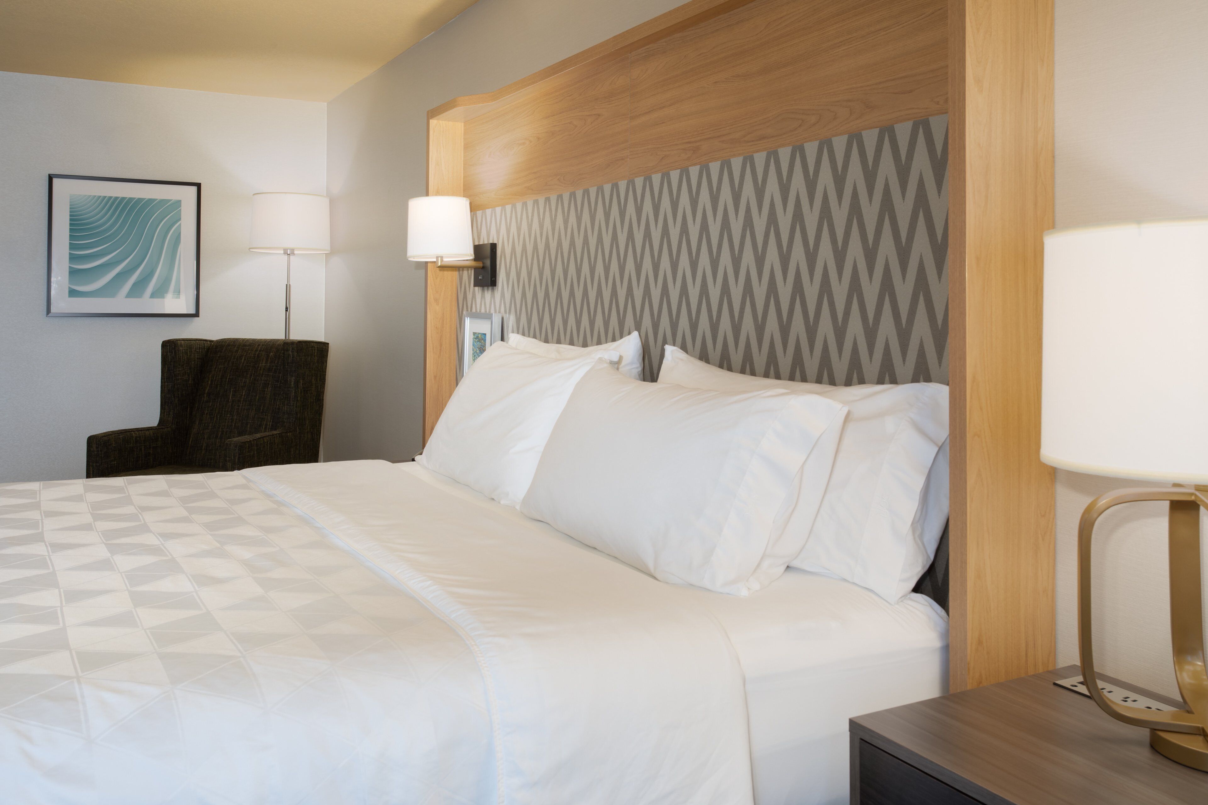 A hotel room with a bed, a chair, a lamp, and a wall-mounted picture.