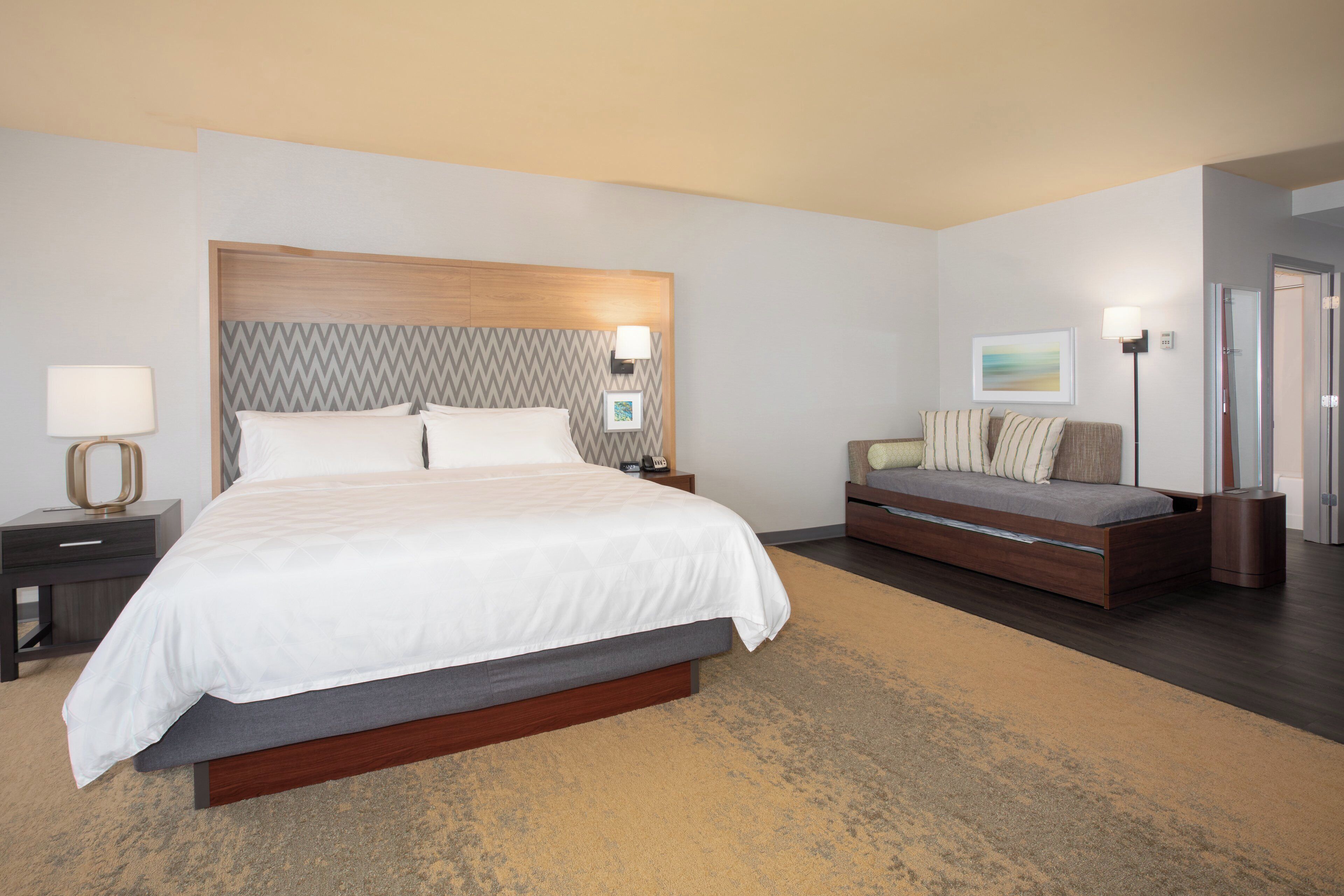 A hotel room with a large bed, a sofa, a bedside table, a lamp, and a painting on the wall.